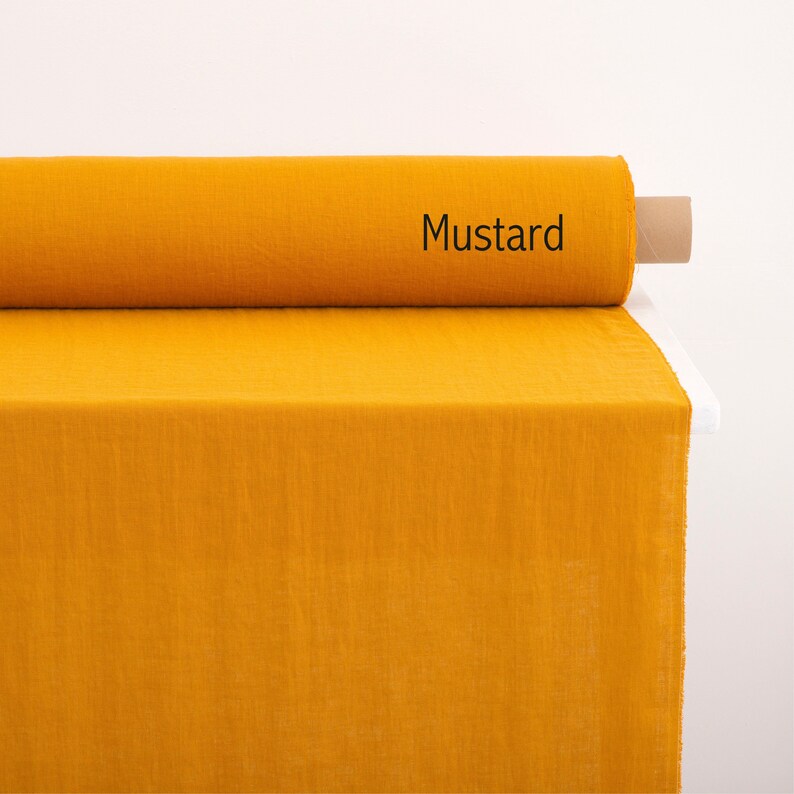 May include: A roll of mustard yellow fabric, unfurled on a white surface. The fabric is a linen blend and has a slightly textured surface. The label on the roll reads "Mustard".