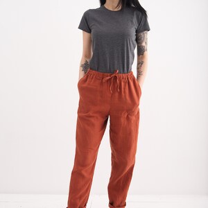 May include: A woman wearing a gray t-shirt and rust-colored linen pants with a drawstring waist. She is wearing beige shoes.