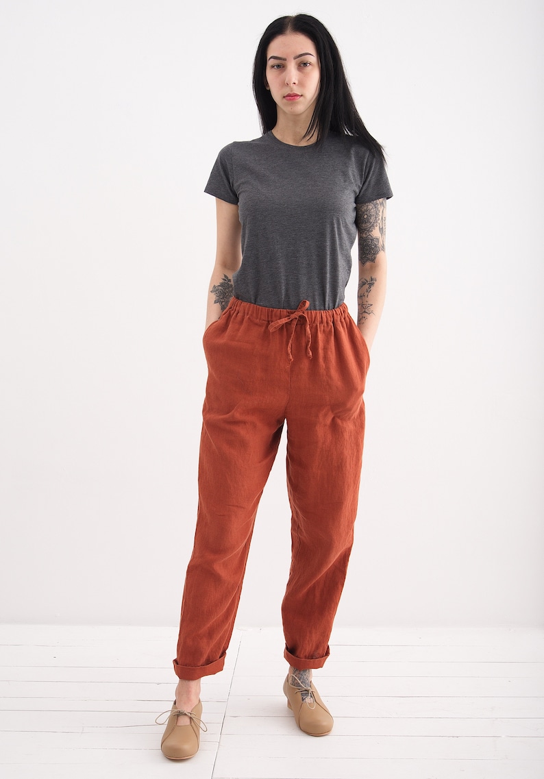 May include: A woman wearing a gray t-shirt and rust-colored linen pants with a drawstring waist. She is wearing beige shoes.