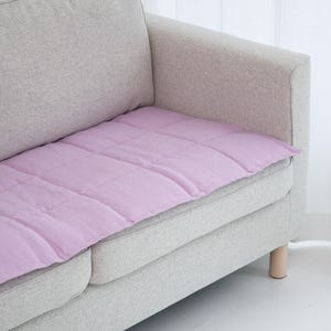 May include: A light gray sofa with a lavender quilted cushion. The cushion is rectangular and covers the seat of the sofa. The sofa has light wooden legs and is placed in front of a white curtain.