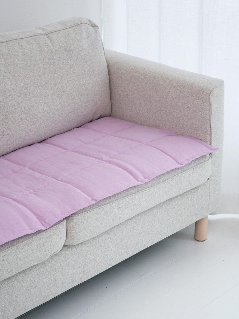 May include: A light gray sofa with a lavender quilted cushion. The cushion is rectangular and covers the seat of the sofa. The sofa has light wooden legs and is placed in front of a white curtain.