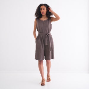 May include: A woman wearing a sleeveless, brown linen jumpsuit with a self-tie belt. She is standing in a white room and looking at the camera.