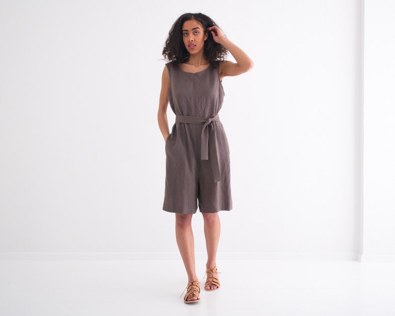 May include: A woman wearing a sleeveless, brown linen jumpsuit with a self-tie belt. She is standing in a white room and looking at the camera.