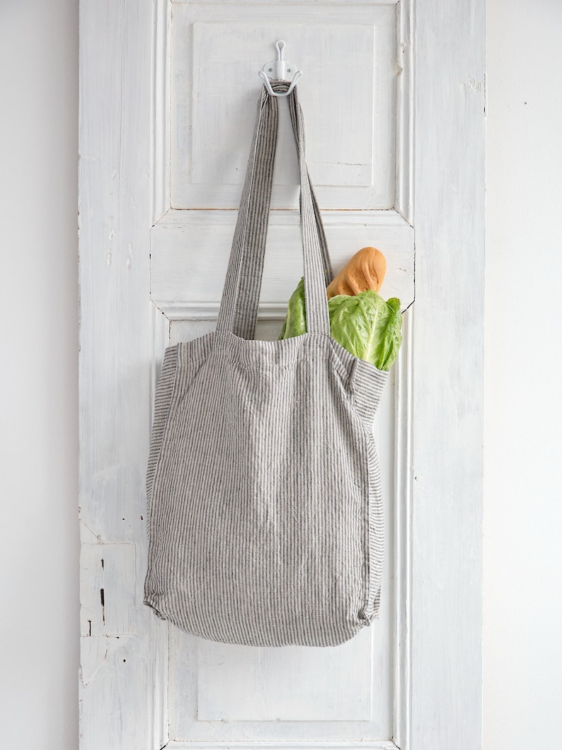 May include: A striped gray and white tote bag hanging on a white door hook. The bag is filled with a loaf of bread and a head of lettuce.