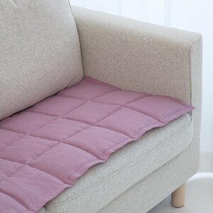 May include: A lavender-colored, quilted cushion placed on a light gray sofa. The cushion has a grid pattern created by stitched lines. The sofa has a light wood leg and is positioned near a white curtain.