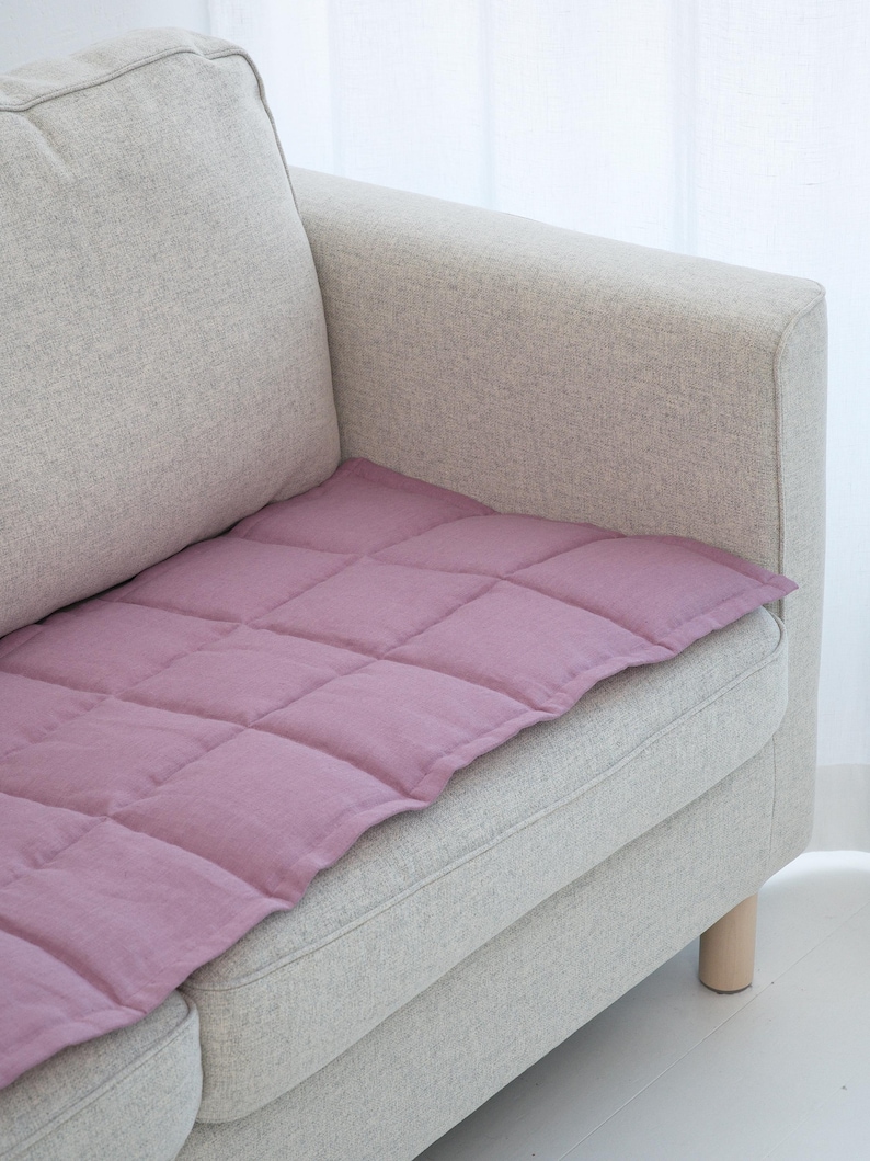 May include: A lavender-colored, quilted cushion placed on a light gray sofa. The cushion has a grid pattern created by stitched lines. The sofa has a light wood leg and is positioned near a white curtain.