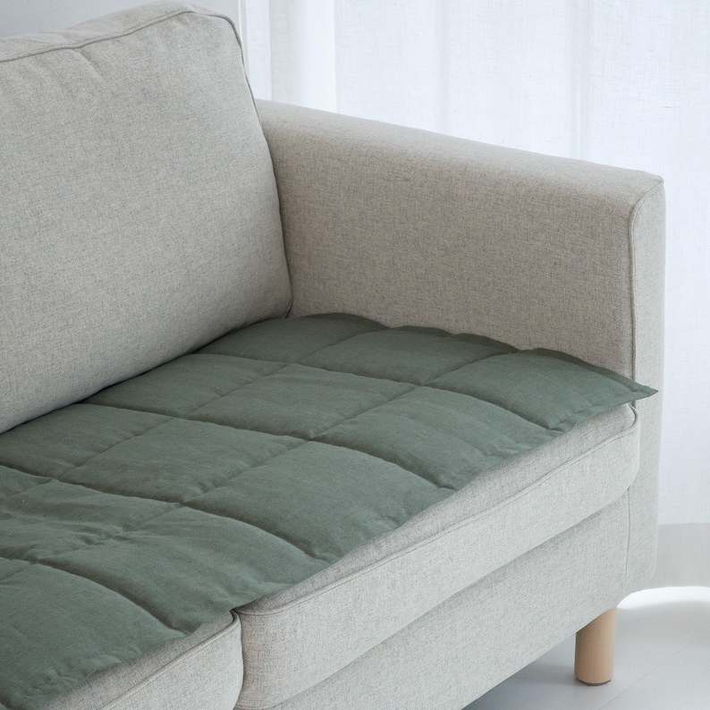 May include: A sage green, quilted cushion placed on the seat of a light gray sofa. The cushion is rectangular and has a grid pattern of stitched squares. The sofa has a neutral color palette and a minimalist design.
