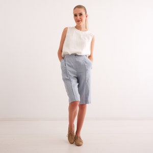 May include: A woman wearing a white sleeveless top and light blue knee-length shorts. The shorts have a high waist and a button closure. The woman is wearing tan shoes. The background is a plain white wall and floor.