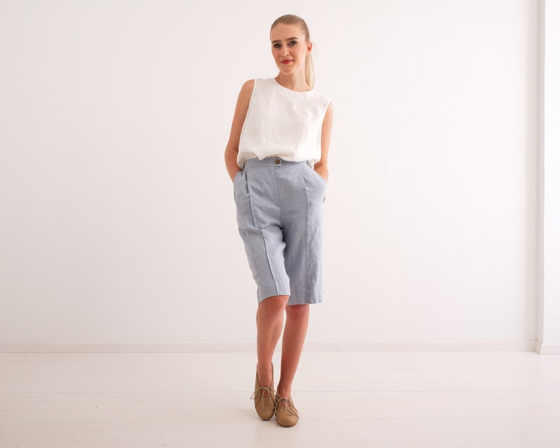 May include: A woman wearing a white sleeveless top and light blue knee-length shorts. The shorts have a high waist and a button closure. The woman is wearing tan shoes. The background is a plain white wall and floor.