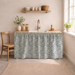 May include: A kitchen sink area with a light wood countertop and a white sink. A patterned fabric skirt with a floral design covers the cabinet area. A wooden chair and a beige rug are in the foreground.