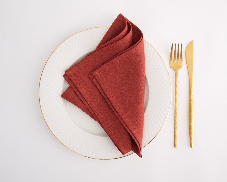 May include: A rust-colored linen napkin folded into a triangle shape is placed on a white plate with a gold rim. A gold fork and knife are placed to the right of the plate.