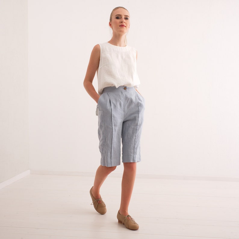 May include: A woman wearing a white sleeveless top and light blue linen shorts. The shorts have a button closure and pockets. She is wearing brown leather shoes. The outfit is simple and elegant, suitable for warm weather.