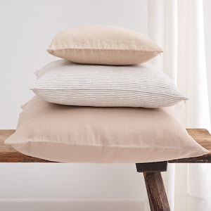 Stack of three deocorative square pillow covers, in the colors beige and striped white with a hidden zipper closure on a wooden bench.