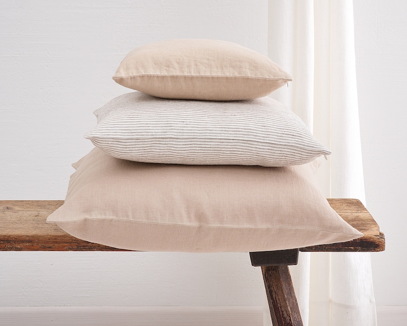 Stack of three deocorative square pillow covers, in the colors beige and striped white with a hidden zipper closure on a wooden bench.