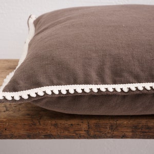 May include: A brown linen cushion with a white pom-pom trim.