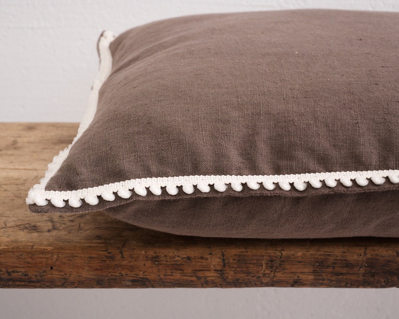May include: A brown linen cushion with a white pom-pom trim.