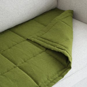 May include: A green, quilted blanket draped over a light gray sofa. The blanket has a square pattern and is made of a textured fabric. The color is a vibrant, earthy green, contrasting with the neutral sofa.