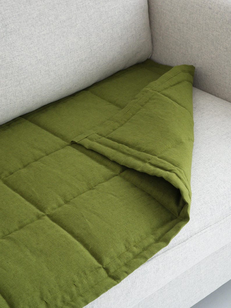 May include: A green, quilted blanket draped over a light gray sofa. The blanket has a square pattern and is made of a textured fabric. The color is a vibrant, earthy green, contrasting with the neutral sofa.