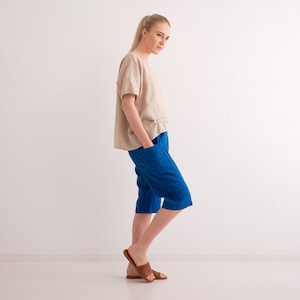 May include: A woman wearing a beige linen top and blue capri pants. The top has short sleeves and a ruffled detail at the waist. The capri pants are a vibrant blue and have pockets. The woman is also wearing brown leather sandals.