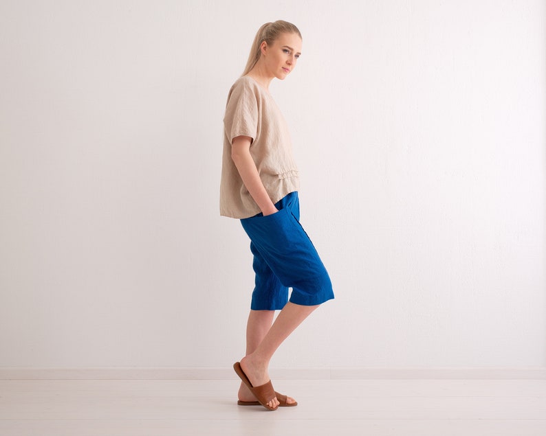 May include: A woman wearing a beige linen top and blue capri pants. The top has short sleeves and a ruffled detail at the waist. The capri pants are a vibrant blue and have pockets. The woman is also wearing brown leather sandals.