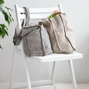May include: Two linen tote bags, one striped and one solid, are sitting on a white folding chair. The striped bag has a brown and white stripe pattern. The solid bag is a light beige color.