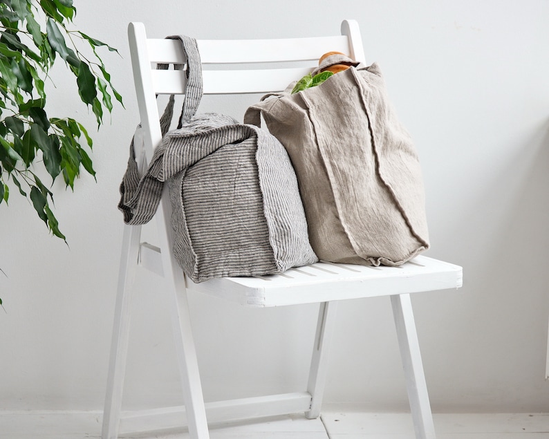 May include: Two linen tote bags, one striped and one solid, are sitting on a white folding chair. The striped bag has a brown and white stripe pattern. The solid bag is a light beige color.