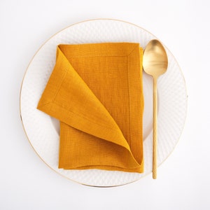 May include: A white plate with a gold rim, a folded mustard yellow linen napkin, and a gold spoon.