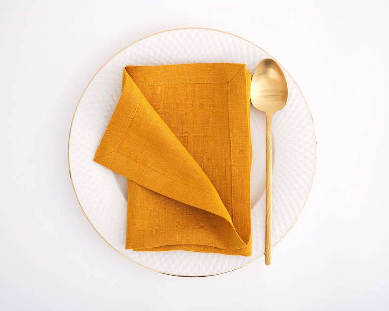 May include: A white plate with a gold rim, a folded mustard yellow linen napkin, and a gold spoon.