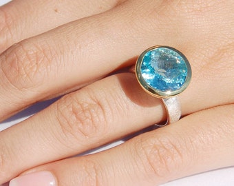 May include: A ring with a large, round, blue gemstone set in a gold bezel. The ring band is made of silver and has a textured finish.