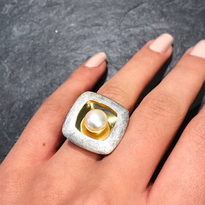 May include: A silver ring with a gold square inset featuring a white pearl. The ring is on a hand with painted nails.