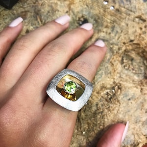 May include: A silver ring with a square design featuring a green gemstone set in a gold center.