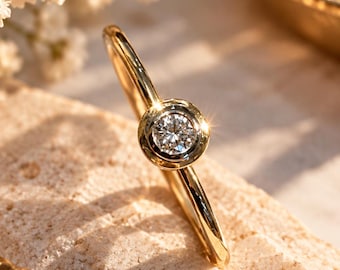 May include: A gold ring featuring a single round brilliant-cut diamond set in a smooth bezel setting. The band is polished yellow gold, and the diamond is positioned to catch the light, creating a bright sparkle against the metal.