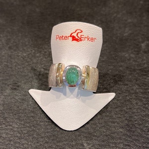 Op de afbeelding: Een zilveren ring met een groene edelsteen gezet in een gouden band. De ring ligt op een witte displaystandaard met de tekst "Peter Erker" in rood.