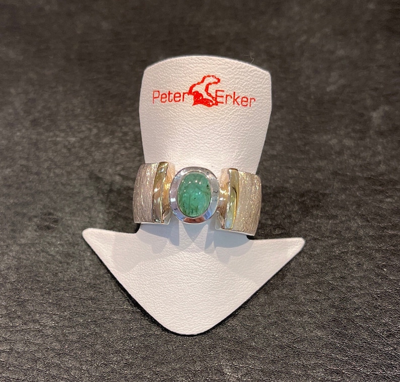 Op de afbeelding: Een zilveren ring met een groene edelsteen gezet in een gouden band. De ring ligt op een witte displaystandaard met de tekst "Peter Erker" in rood.