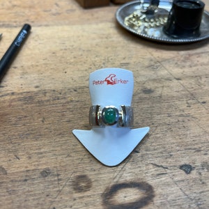 Op de afbeelding: Een zilveren ring met een groene edelsteen gezet in een zilveren band. De ring ligt op een witte displaystandaard met de tekst "Peter Erker" erop gedrukt.