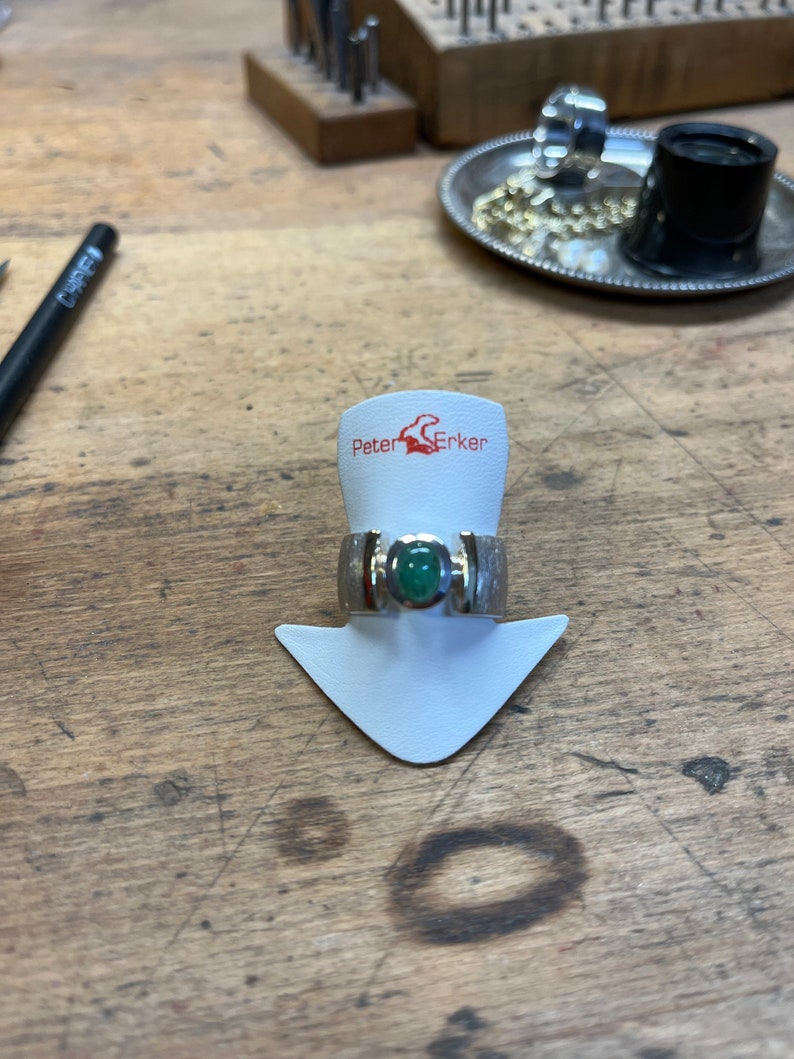 Op de afbeelding: Een zilveren ring met een groene edelsteen gezet in een zilveren band. De ring ligt op een witte displaystandaard met de tekst "Peter Erker" erop gedrukt.