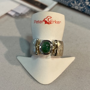 Op de afbeelding: Een zilveren en gouden ring met een groene ovale edelsteen. De ring ligt op een witte displaystandaard met de tekst "Peter Erker" erop.