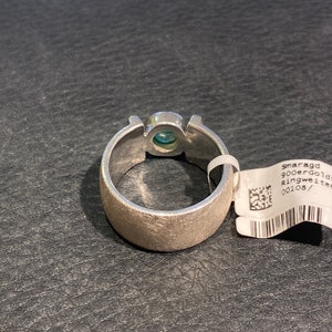 Op de afbeelding: Een zilveren ring met een groene edelsteen in het midden. De ring ligt op een zwart, getextureerd oppervlak. Het label dat aan de ring is bevestigd, zegt "smaragd 900er gold ringweite 00108/"
