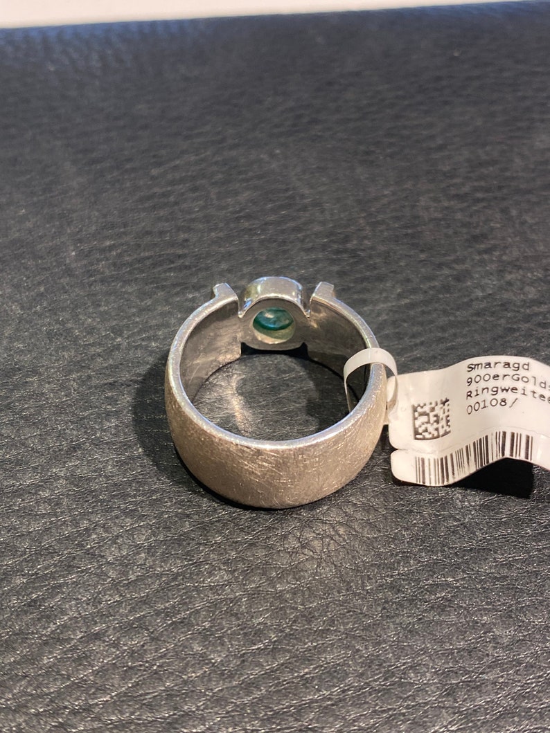 Op de afbeelding: Een zilveren ring met een groene edelsteen in het midden. De ring ligt op een zwart, getextureerd oppervlak. Het label dat aan de ring is bevestigd, zegt "smaragd 900er gold ringweite 00108/"