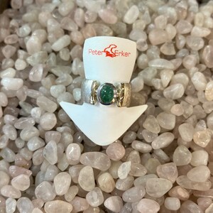 Op de afbeelding: Een zilveren ring met een groene edelsteen gezet in een zilveren band. De ring wordt getoond op een witte standaard met de tekst "Peter Erker" op de standaard.