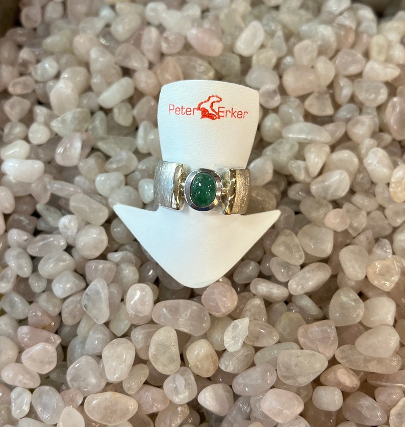Op de afbeelding: Een zilveren ring met een groene edelsteen gezet in een zilveren band. De ring wordt getoond op een witte standaard met de tekst "Peter Erker" op de standaard.