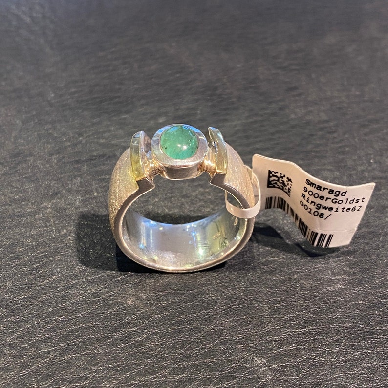 Op de afbeelding: Een zilveren ring met een groene smaragd edelsteen gezet in een gouden band. De ring heeft een geborstelde afwerking en is gelabeld "Smaragd 900erGoldst Ringweite 62 00108/"