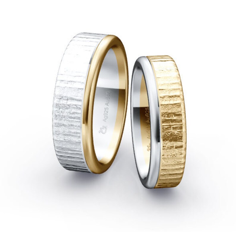 Op de afbeelding: Twee trouwringen, een met een zilveren en gouden afwerking en de andere met een gouden en zilveren afwerking. Beide ringen hebben een getextureerd oppervlak.