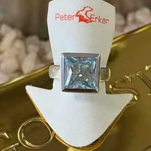 Könnte beinhalten: Ein silberner Ring mit einem quadratischen, hellblauen Edelstein. Der Ring befindet sich auf einem weißen Präsentationsständer mit dem Text "Peter Erker" in Rot.