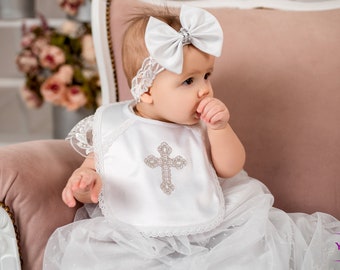 May include: A white baby dress with a lace trim and a white bib with a silver cross design. The baby is wearing a white headband with a large white bow and a silver rhinestone.