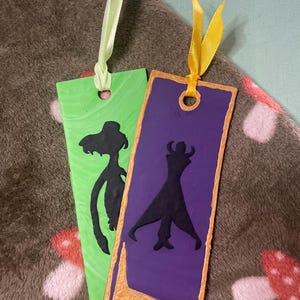 May include: Two handmade bookmarks with colourful backgrounds and black silhouette figures. The bookmark on the left has a green background and a silhouette of a woman. The bookmark on the right has a purple background and a silhouette of a man.