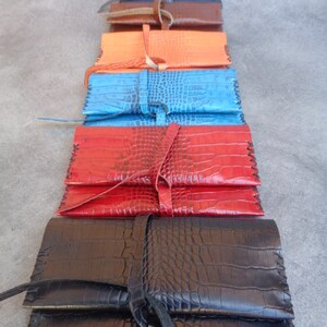 Könnte beinhalten: Ein Stapel von acht Zigarettenetuis aus Leder in verschiedenen Farben, darunter Schwarz, Rot, Blau, Orange, Braun und Dunkelblau. Die Etuis sind aus geprägtem Leder mit Kroko-Muster und haben einen Klappverschluss mit Lederband.