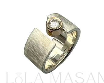 May include: A silver and gold ring with a single round diamond set in a bezel setting. The ring is brushed and has a unique design.