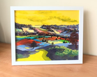 May include: A colorful abstract landscape painting with a yellow sky and green, blue, red, and purple hills. The painting is framed in a white frame.