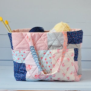 Può includere: Una borsa a mano in tessuto patchwork con un motivo floreale rosa, blu e bianco. La borsa ha un'unica maniglia ed è piena di filato.
